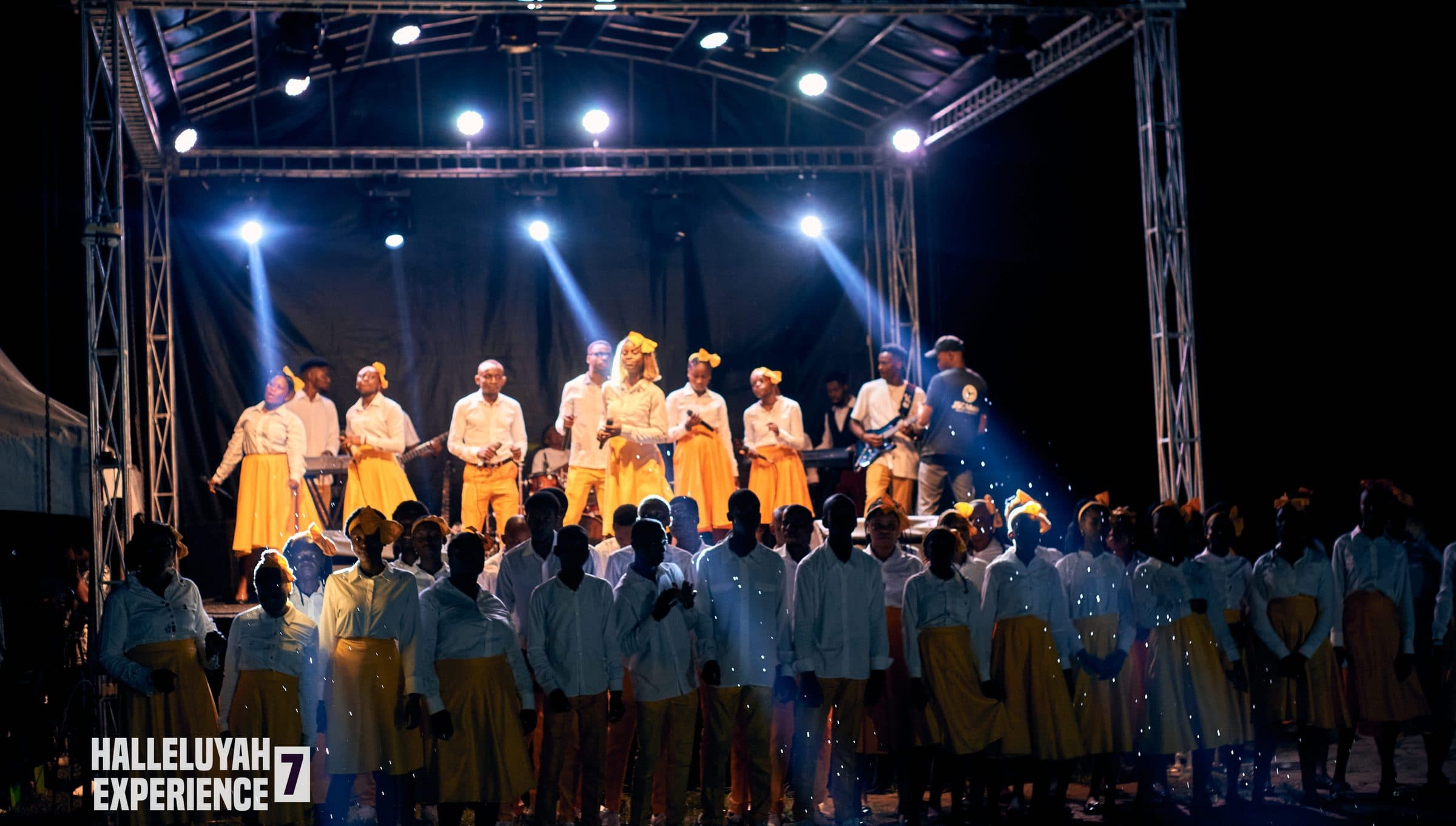 The full choir performing on a lit stage at night, the rig visible overhead.